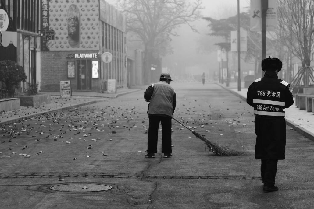 Ein schwarz-weiß Foto, dass eine weitgehend leere Straße im Dunst zeigt, eine Straßenkehrerin fegt Laub zusammen, ein Baoan (Wächter) sieht ihr zu.
