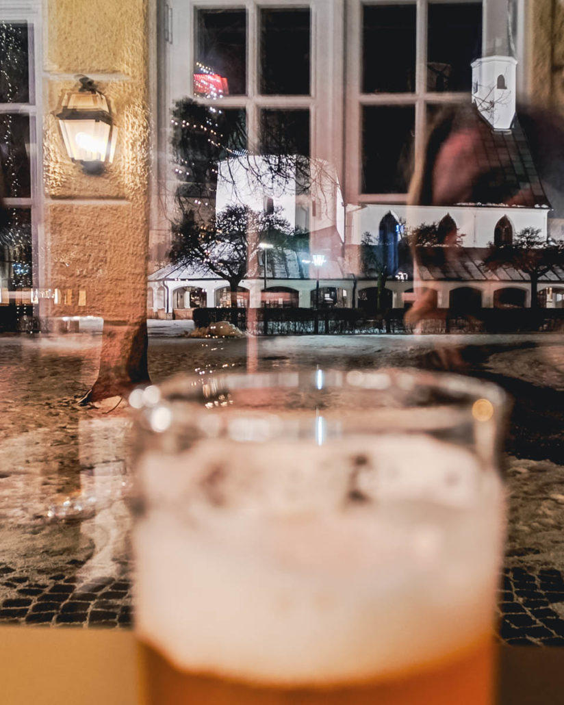 Durch ein Wintergartenfenster fotografierte Gnadenkappel auf dem Kapellplatz in Altötting, im Vordergrund ein Weizenbierglas, Spiegelungen im Fenster